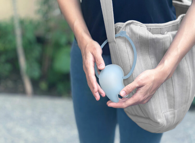 Person holding a light blue portable urinal funnel next to a beige shoulder bag, with an outdoor background visible.