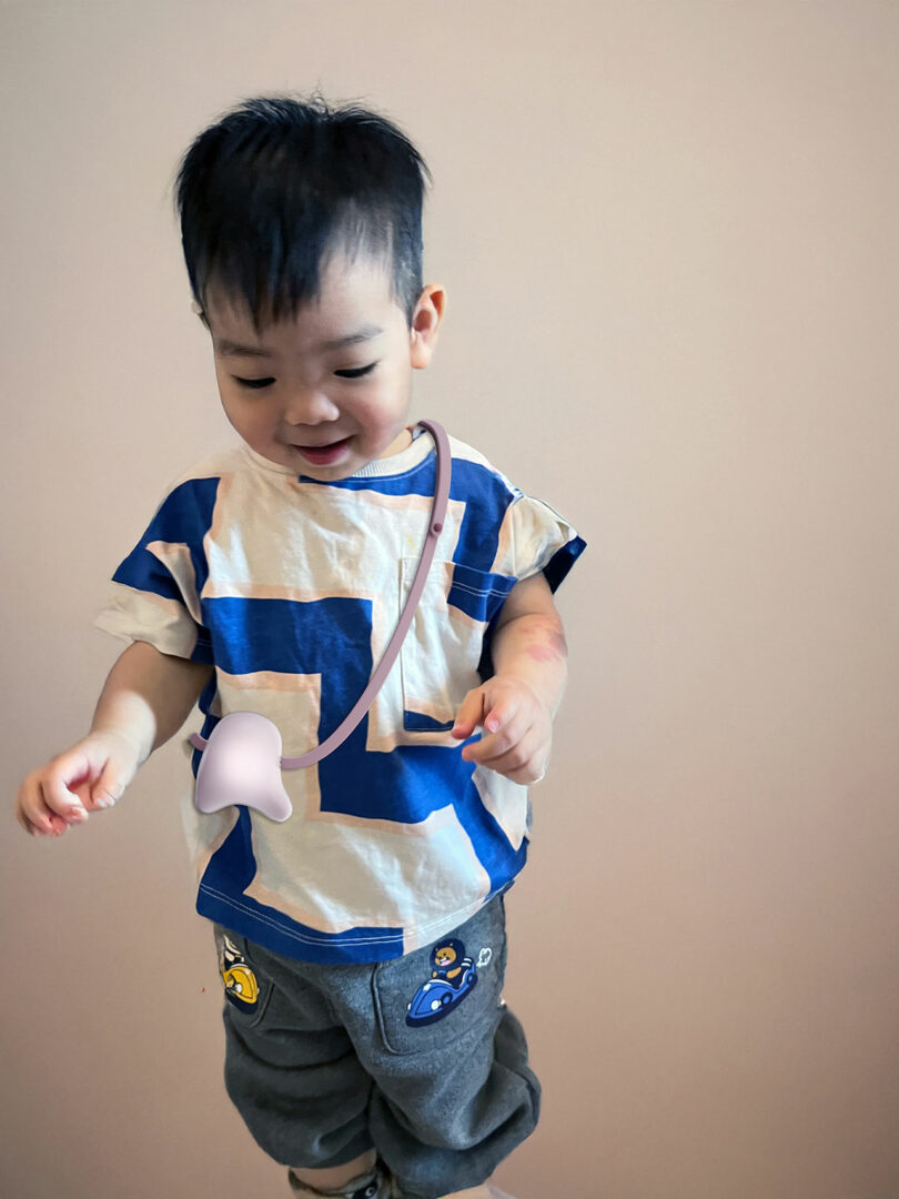 Young child wearing a blue and white shirt and grey pants with patches, standing against a plain light-colored background, with a TruTru Haptic Companion around their neck.