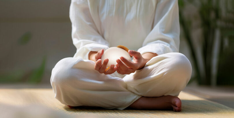 A person in loose white clothing sits cross-legged on a mat, holding a round, glowing object in their hands.
