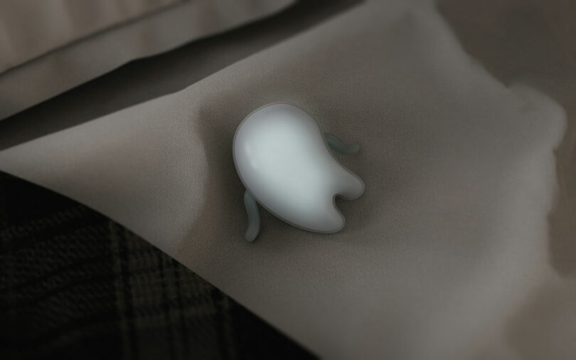 A small, smooth, ghost-shaped object with two short arms lies on a beige fabric surface.