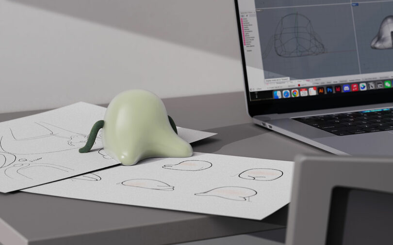 A small, green, blob-like model with two tentacles sits on sketches next to a laptop displaying a 3D modeling program on a grey desk.