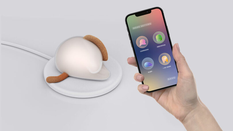 A hand holds a smartphone displaying four app icons near a small, helmet-shaped device with brown side attachments on a circular pad.