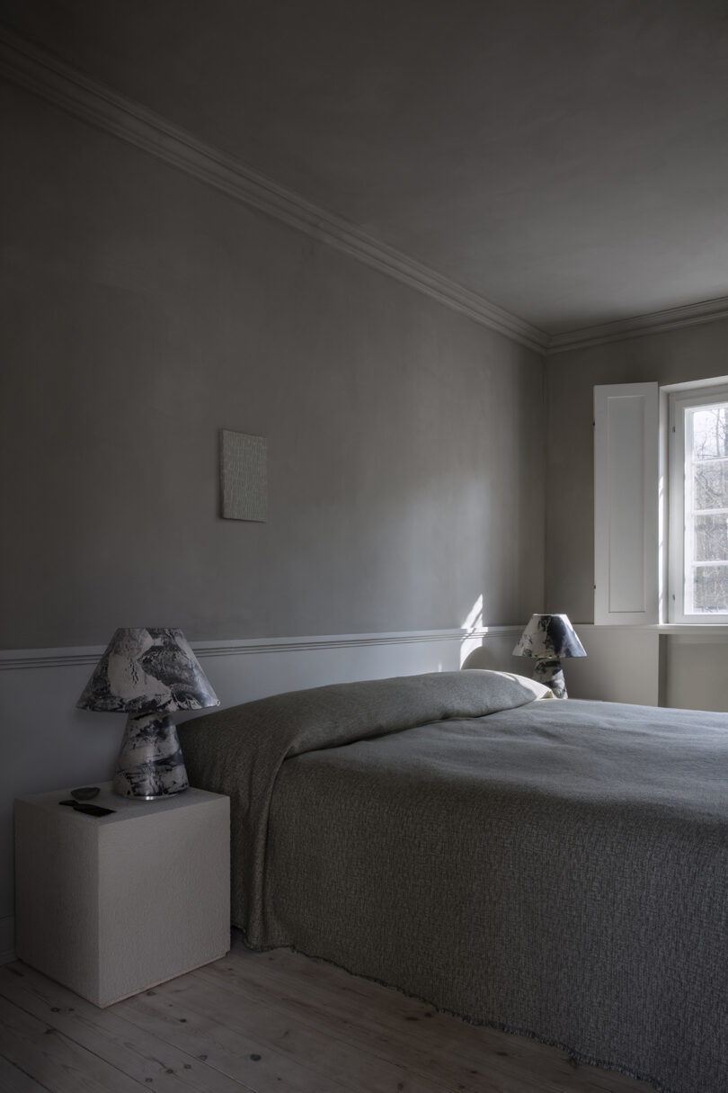 A minimalist bedroom with a neatly made bed, two patterned lamps on either side, light wood floor, and natural light coming in through a window with white shutters.