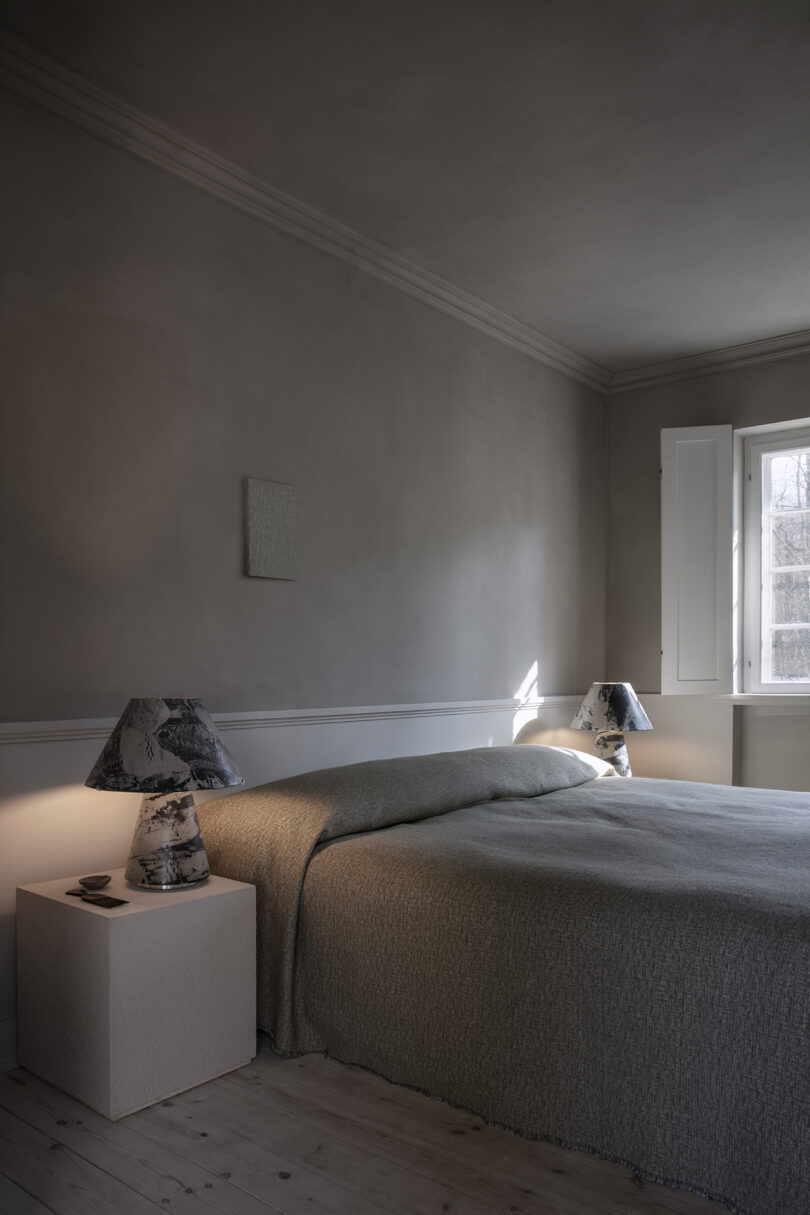 A minimalist bedroom with a neatly made bed, two nightstands, matching lamps, and a window allowing natural light into the space.