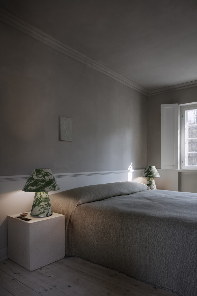 A minimalist bedroom with a neatly made bed, two green marble lamps on side tables, neutral walls, wooden floor, and sunlight coming through a window.