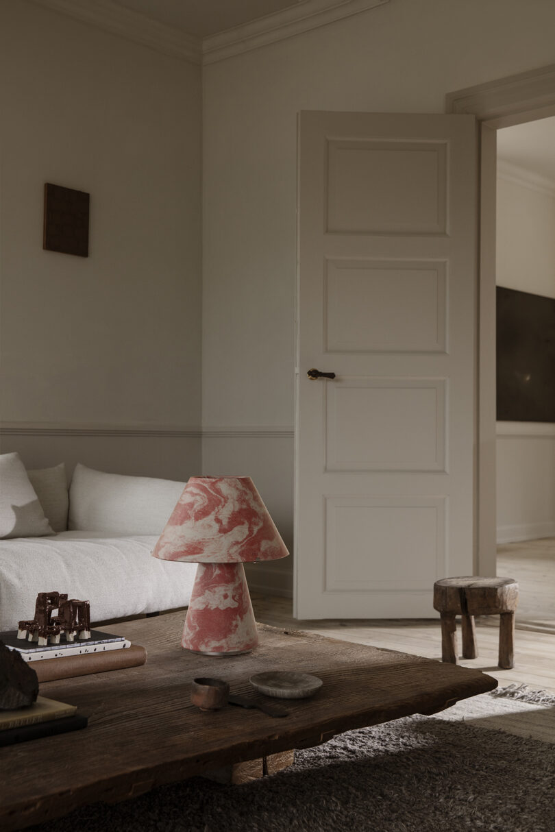 A cozy living room with a white sofa, a wooden coffee table holding books, a pink marbled lamp, small decor items, and an open door leading to another room.