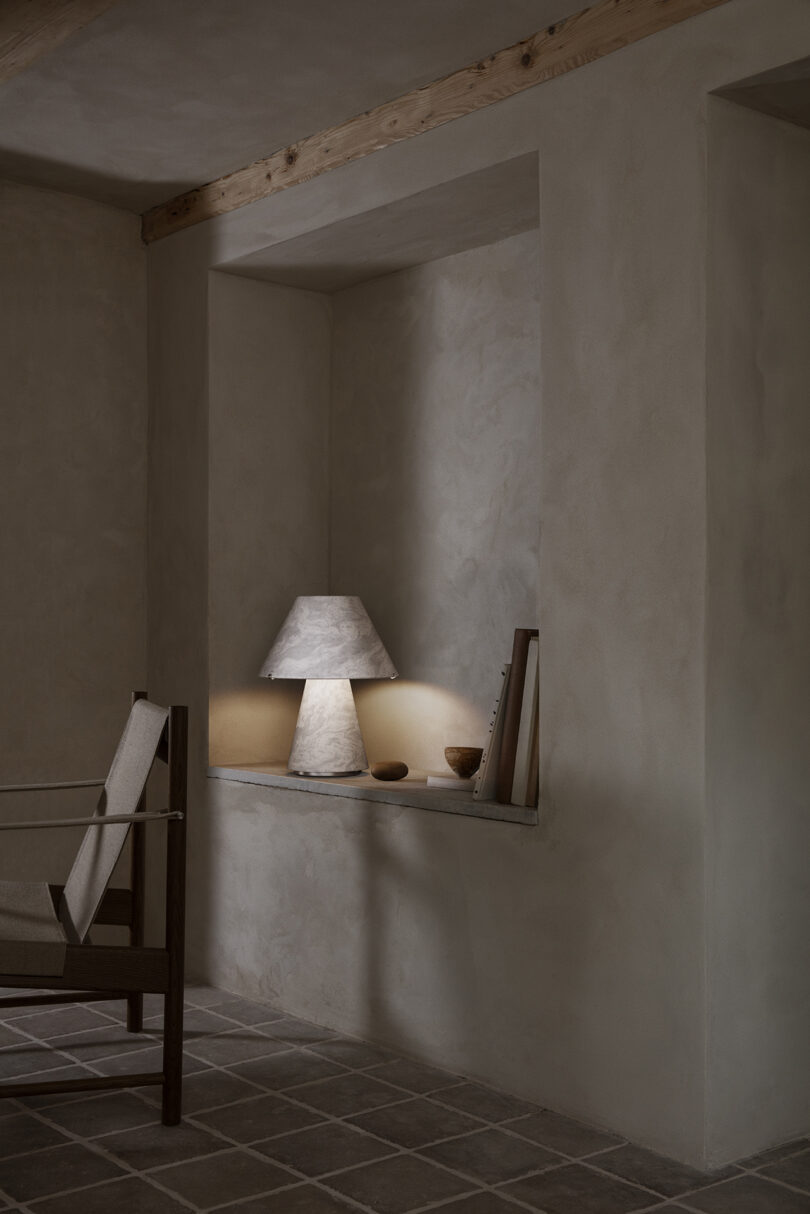 A minimal alcove features a stone table lamp, two small bowls, and several books, with a wooden chair in the foreground on a tiled floor.