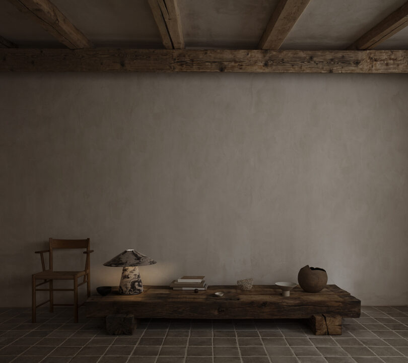 A minimalist room with a wooden ceiling beam, a simple chair, a low rustic coffee table with books, a lamp, pottery, and decorative objects, set against a plain textured wall.