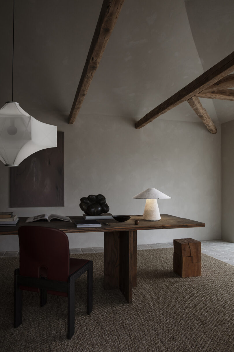 Minimalist workspace with a wooden desk, modern lamp, dark chair, abstract sculpture, and pendant light under exposed wooden beams.
