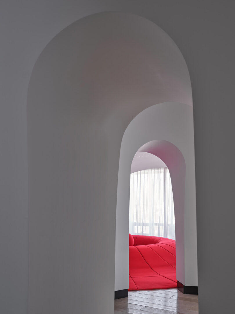 A smooth, white arched doorway frames a bright room with a large red, modern sofa and floor-to-ceiling sheer curtains illuminated by natural light.