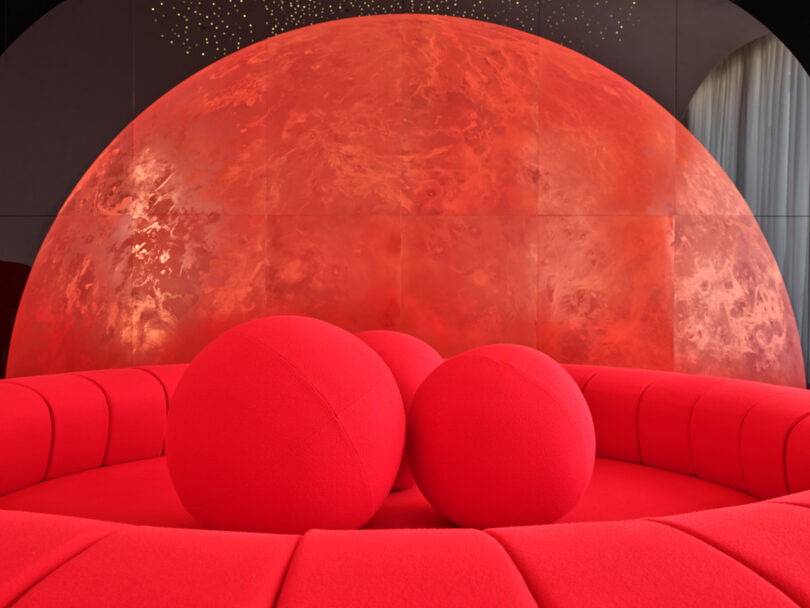 A circular red couch with three large red spheres sits in front of a large, illuminated, semi-circular red backdrop resembling a planet.