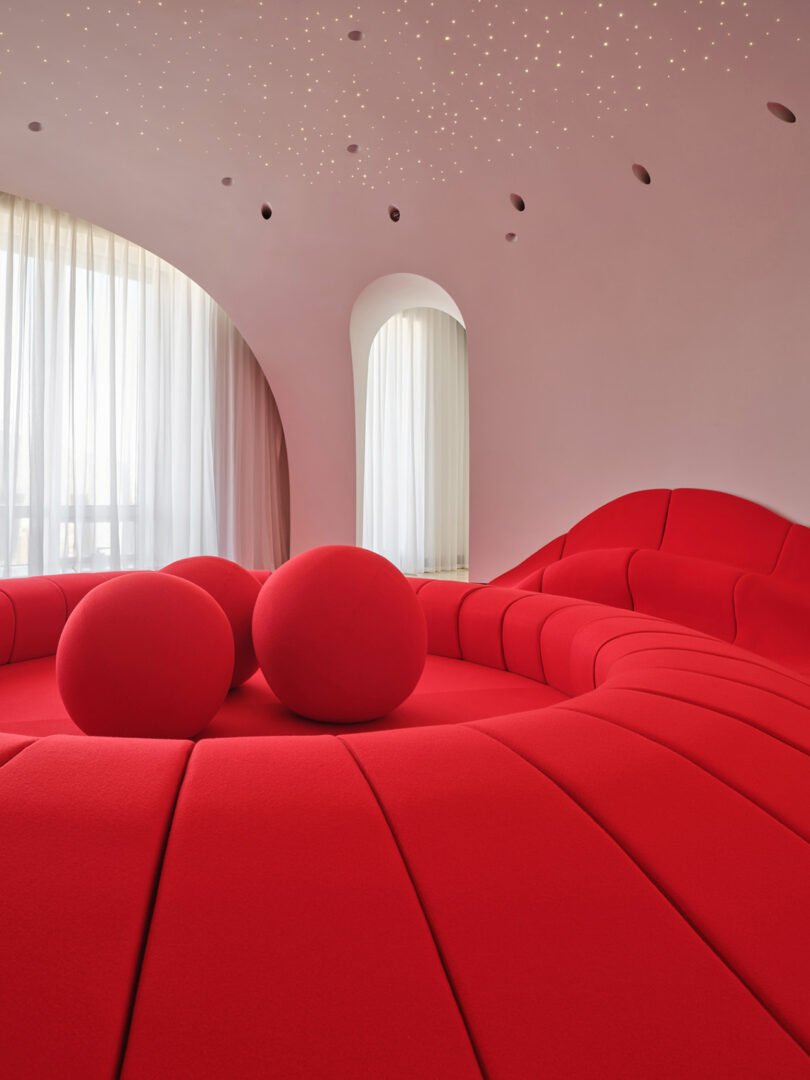 A modern room with a large, curved red sofa and three spherical red cushions, set against white walls with arched windows and a ceiling dotted with small lights.