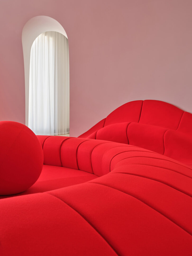 A modern interior with a sculptural, bright red sofa set against pale pink walls, next to an arched window with sheer white curtains.