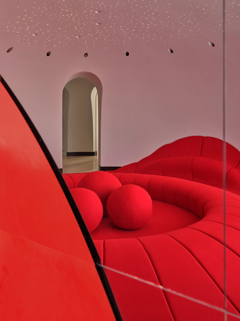 A modern room with a curved red sectional sofa and spherical cushions, a rounded doorway, and a ceiling with small star-like lights.