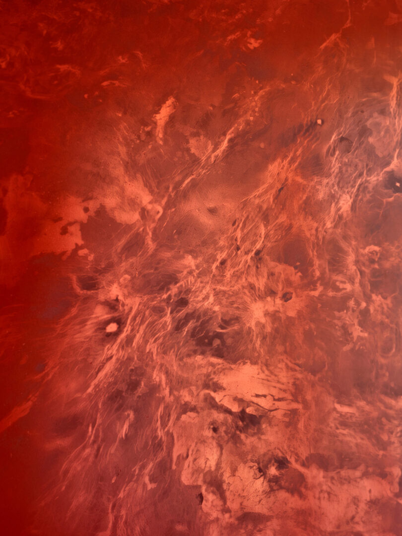 An abstract image featuring swirling shades of red, orange, and pink resembling clouds or smoke on a textured surface.