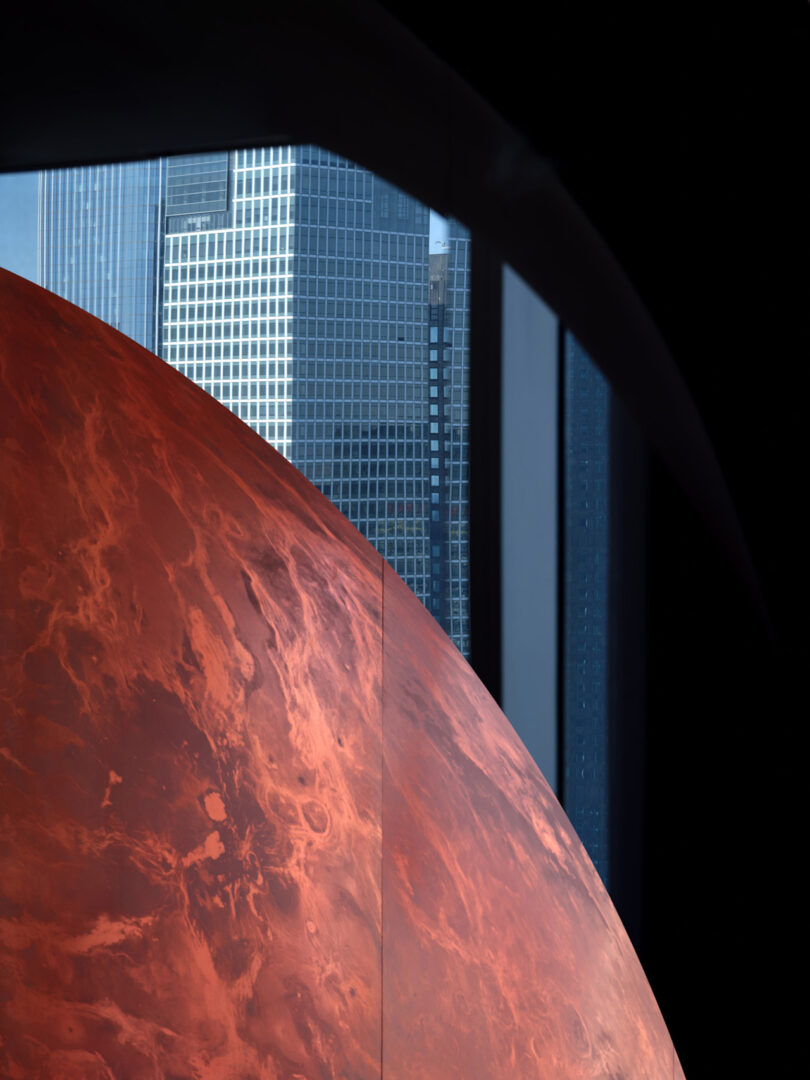 A large, reddish sphere partially in view indoors, with modern city skyscrapers visible through a nearby window.