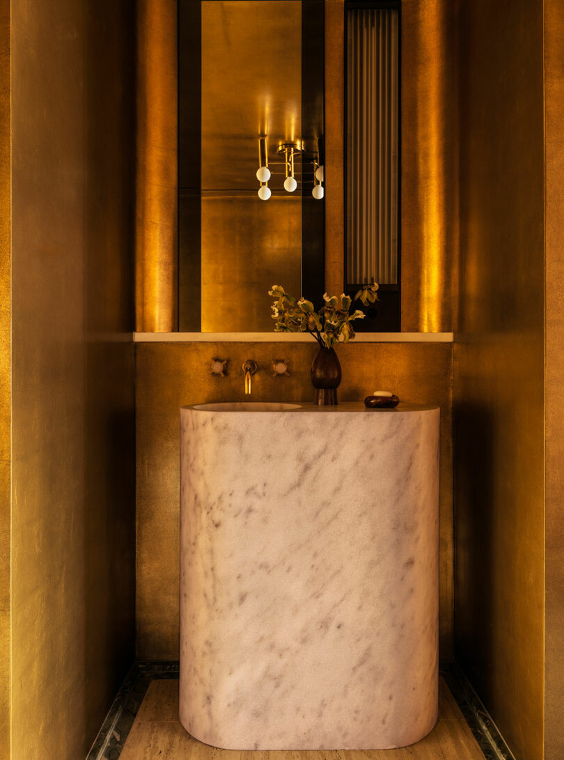 A narrow bathroom features gold walls, a large rectangular mirror, a curved marble sink, a modern light fixture, and a small vase with flowers on the counter.