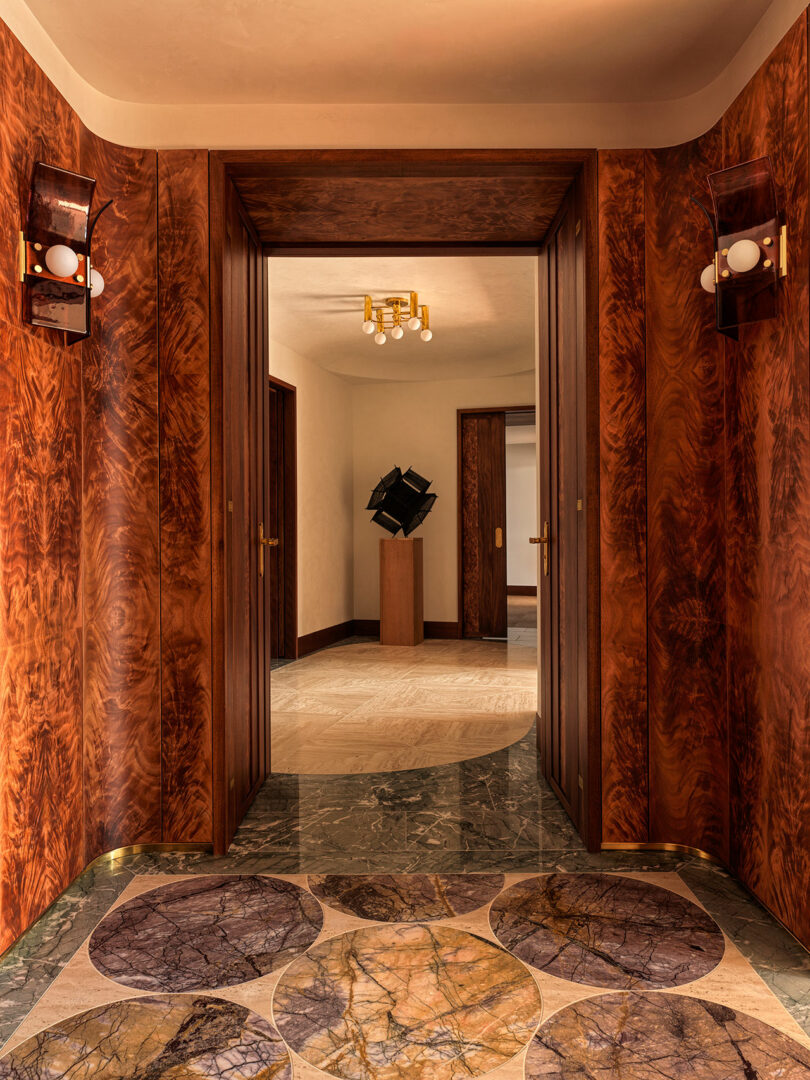 A hallway with wood-paneled walls, marble flooring with circular patterns, and a view into a room featuring a geometric black sculpture on a pedestal.