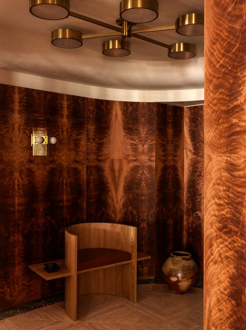 A small seating area with curved, wood-paneled walls, a wooden bench with a cushion, a ceramic pot, and a modern brass ceiling light fixture.