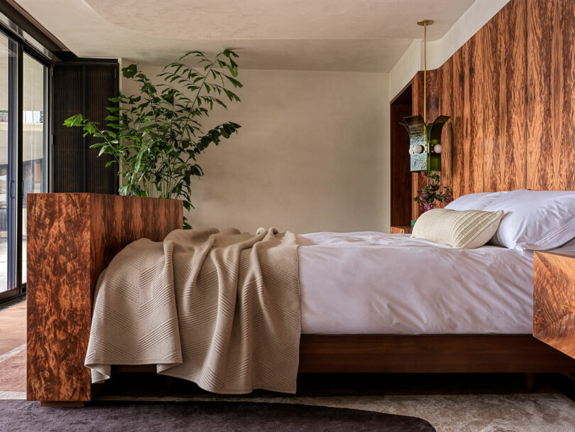 A modern bedroom with a wooden bed frame, neatly made white bedding, a beige blanket, a green plant, and wood-paneled walls.