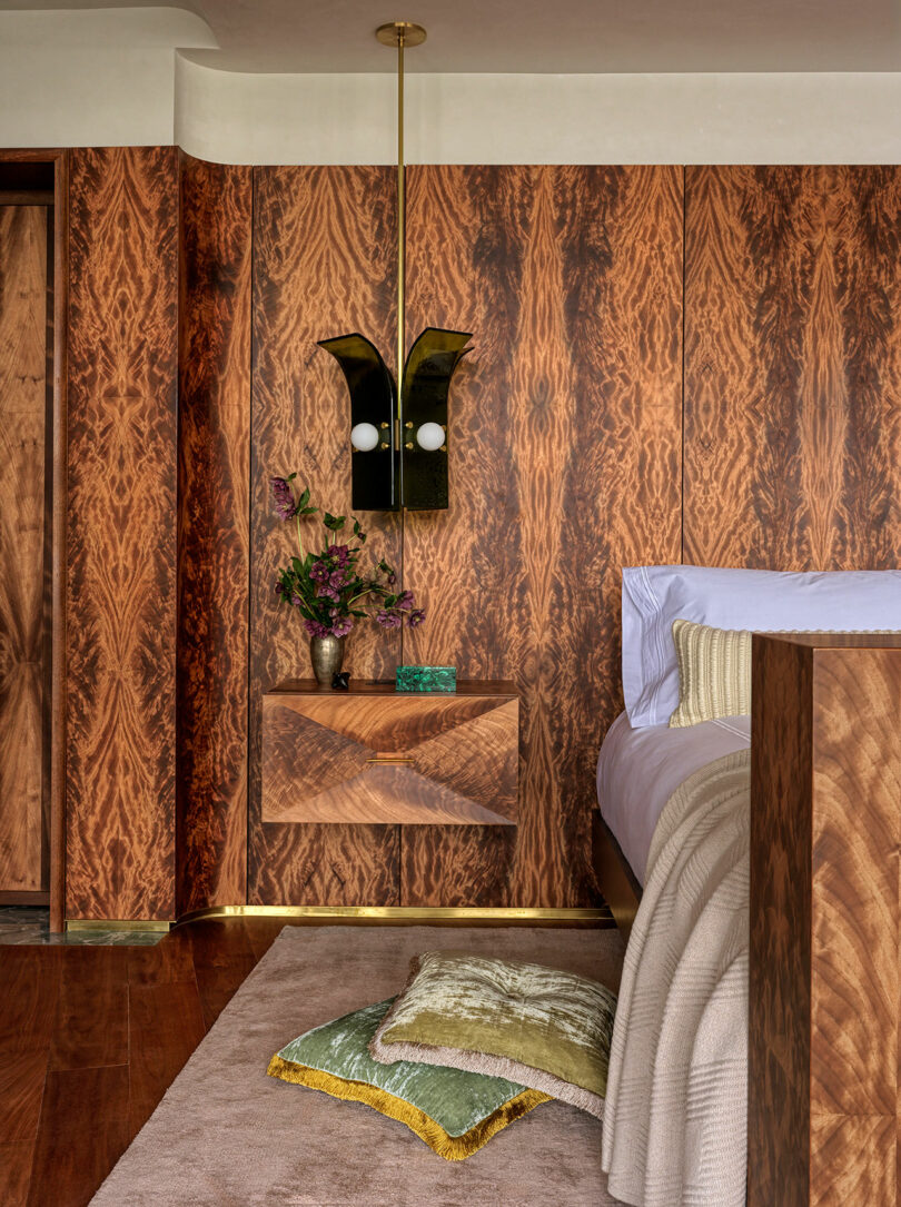A bedroom corner with wood-paneled walls, a floating nightstand, a brass wall sconce, a vase with flowers, and decorative cushions on a beige rug beside the bed.