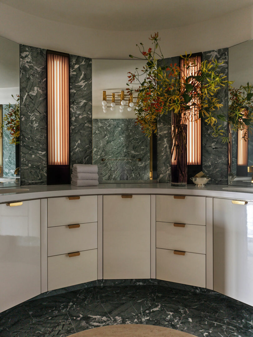 Curved bathroom vanity with white cabinets, brass hardware, green marble surfaces, tall mirrors, wall lights, folded towels, and floral arrangements.