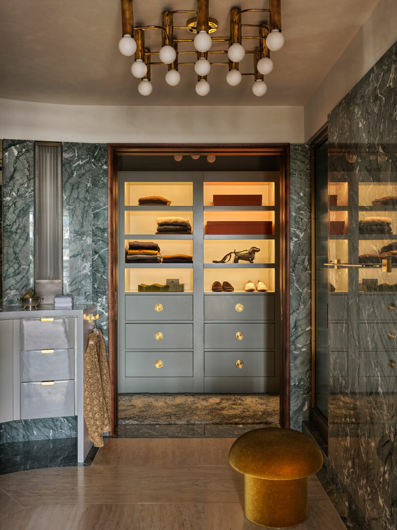 A modern walk-in closet with illuminated shelves, folded clothes, drawers with gold handles, marble walls, and a gold velvet stool in the foreground.