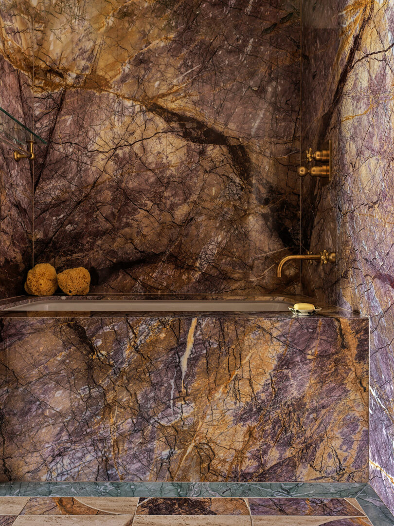 Luxurious bathroom with a bathtub, walls, and floors covered in multicolored marble. Brass fixtures and two natural sponges rest on the tub edge.