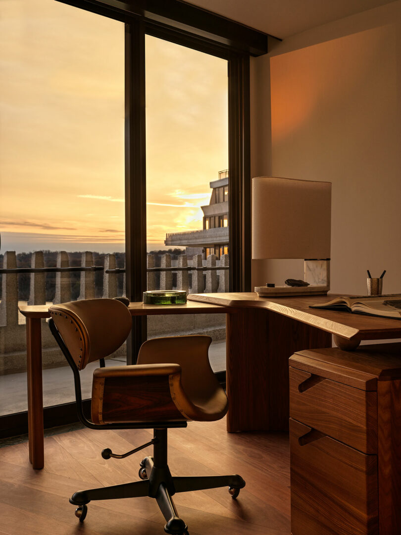 A wooden desk and chair are positioned by large windows overlooking a balcony at sunset, with a lamp, a book, and a pen placed on the desk.