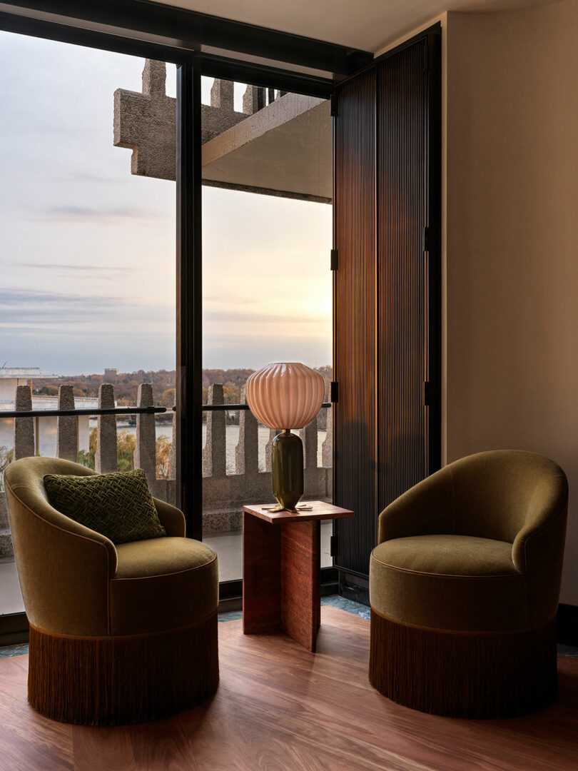 Two olive-green armchairs and a small table with a sculptural lamp are arranged by a window overlooking a balcony with a scenic outdoor view at sunset.