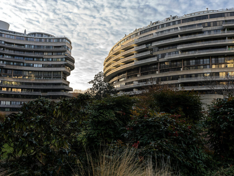 Two curved, modern apartment buildings with balconies rise behind dense greenery under a partly cloudy sky.