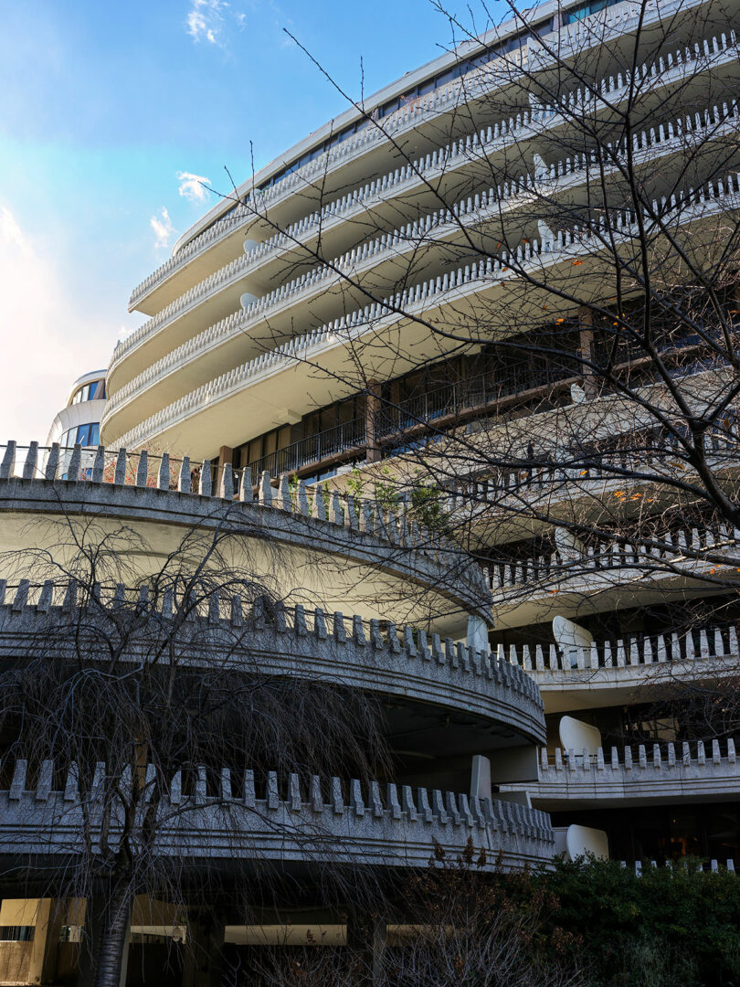 Curved, multi-story concrete building with distinctive repeating vertical elements on balconies and leafless trees in the foreground.