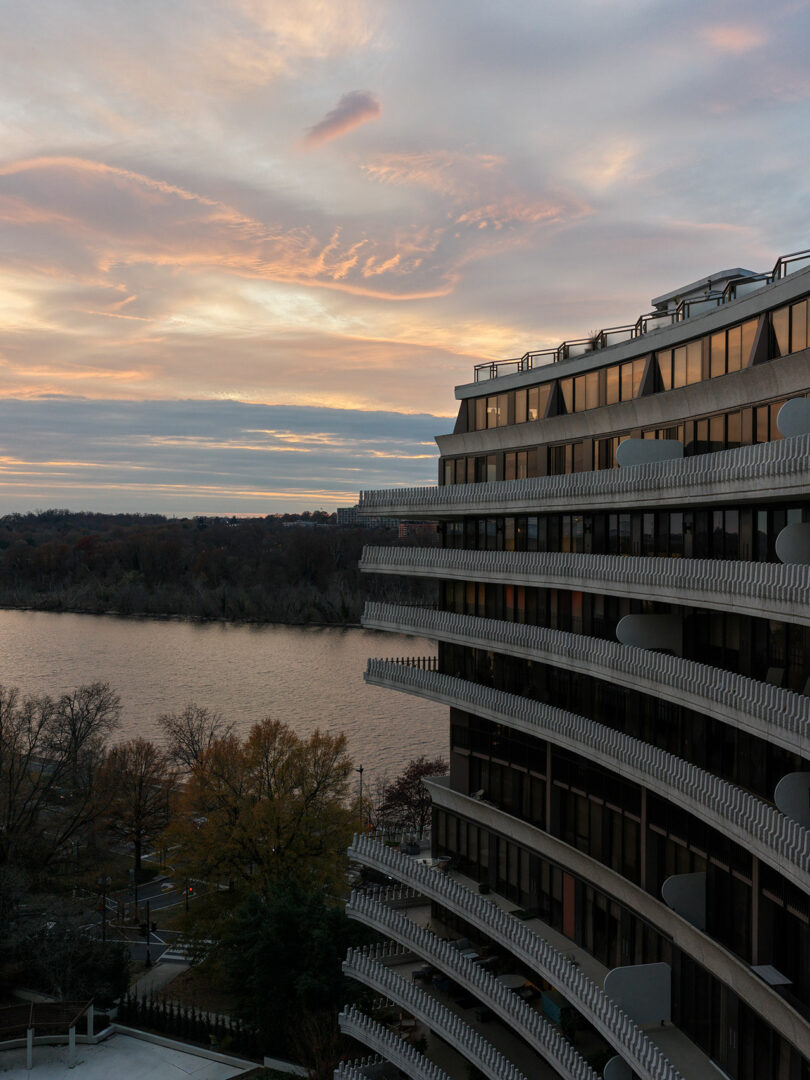 A modern, tiered building overlooks a river with leafless trees on the far bank and a cloudy sunset sky in the background.