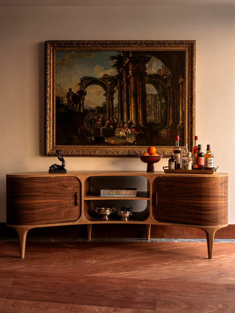 A wooden sideboard with drinks and fruit sits below a framed classical painting on a wall, with silverware and books displayed on open shelves.
