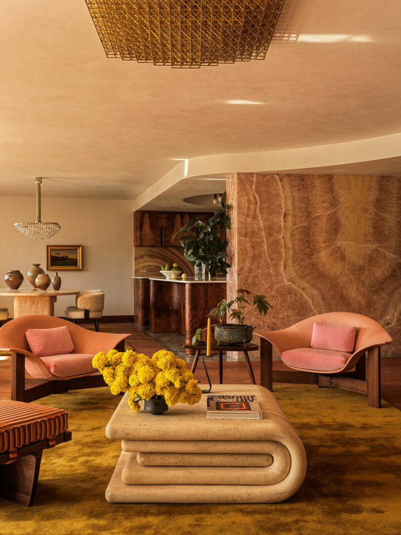 A living room with retro-style furniture, yellow rug, pink chairs, a stone coffee table with books and yellow flowers, and a textured stone wall in the background.
