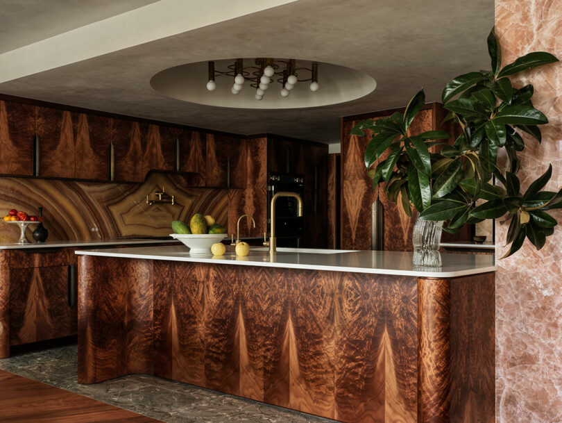 Modern kitchen with glossy wood cabinets, a large island with a white countertop, a decorative plant in a glass vase, and a ceiling light fixture. Fruit bowls sit on the counter.