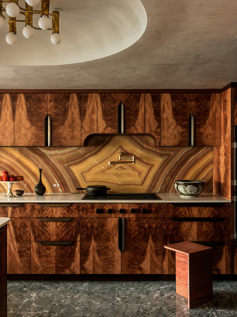 Modern kitchen with patterned wood cabinets, stone backsplash and countertop, gold pot filler, marble floor, and a decorative bowl on the counter.