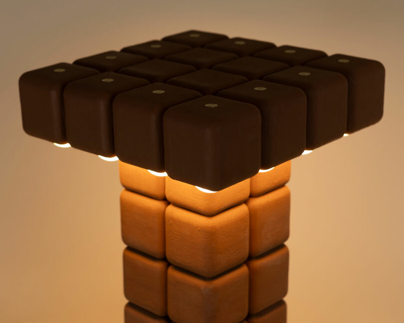 A lamp with a square grid of brown cube shapes by Winkle Ceramic Design emits light from beneath the cubes, creating a warm, ambient glow.