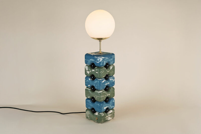 A table lamp with a round white bulb on top of a stacked ceramic base featuring wavy blue and green sections in the signature Winkle Ceramic Design, finished with a visible black power cord.
