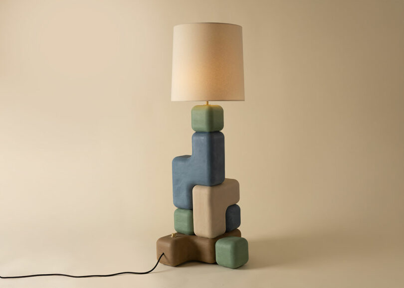 A floor lamp with a beige shade and a base featuring stacked, geometric Winkle Ceramic Design shapes in muted blue, green, brown, and beige tones. A power cord extends from the base.