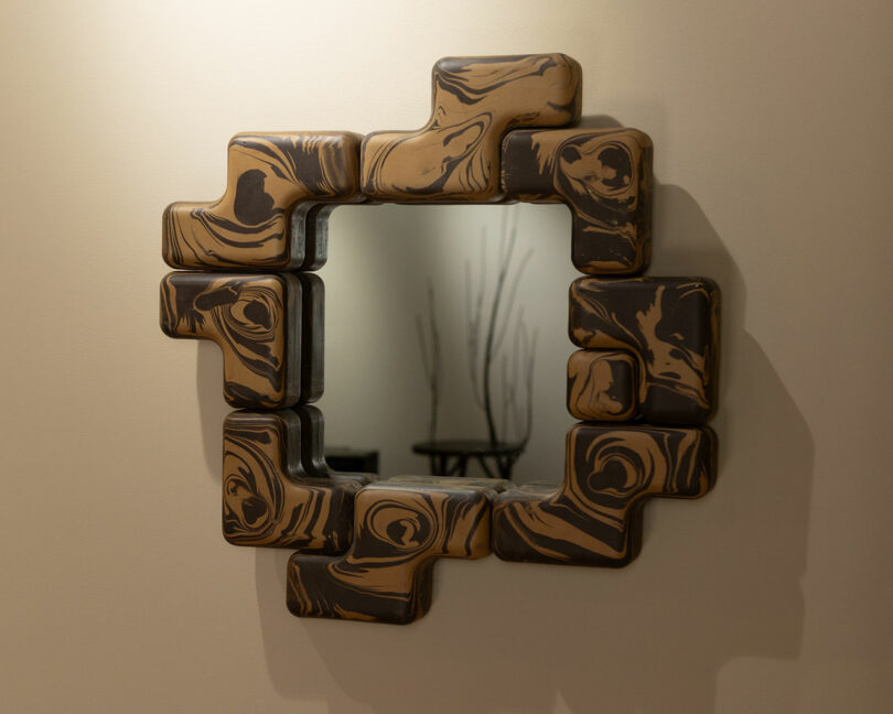 A square mirror with an abstract, geometric wooden frame featuring brown and tan swirled patterns by Winkle Ceramic Design is mounted on a beige wall.