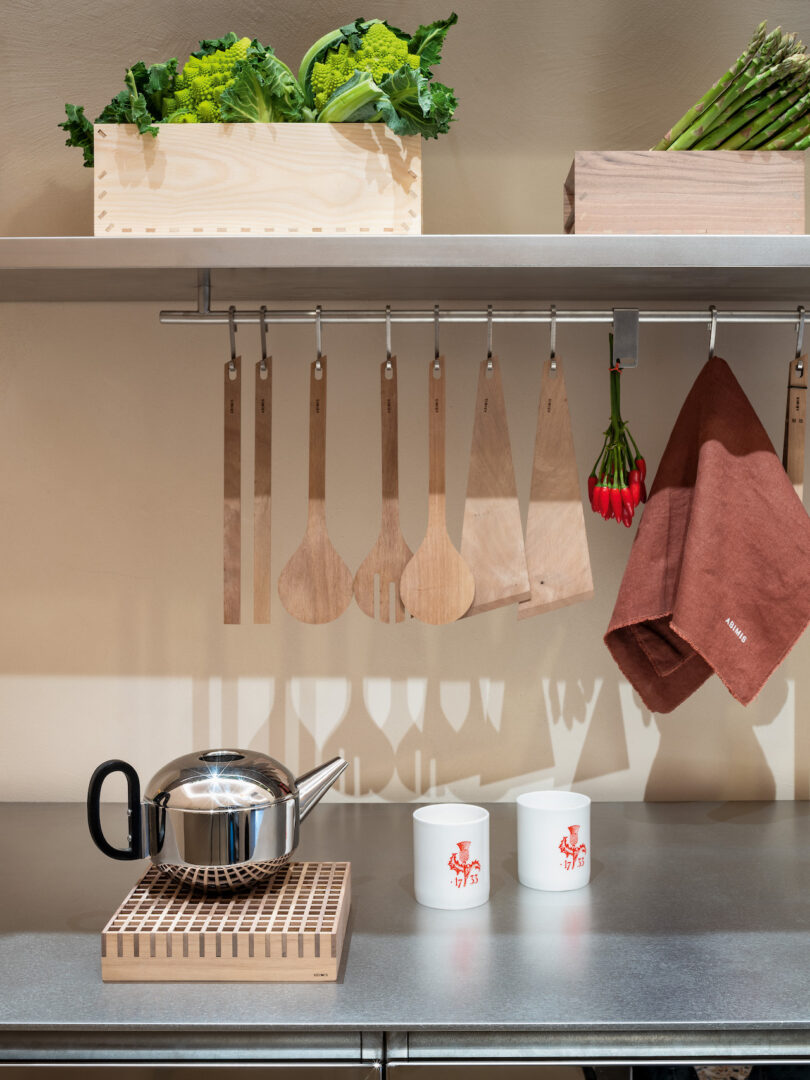A stainless steel kitchen counter with a teapot, two white mugs, wooden utensils hanging above, red chili peppers, towels, and a crate of fresh vegetables on the upper shelf