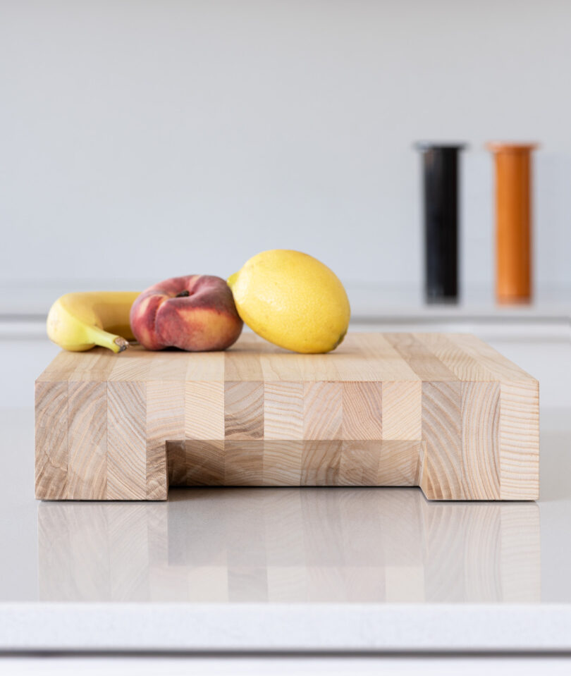 A banana, a peach, and a lemon sit on a wooden cutting board on a kitchen counter with two containers in the background