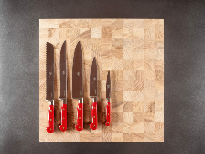 Five kitchen knives with red handles are arranged in descending order of size on a wooden cutting board, viewed from above