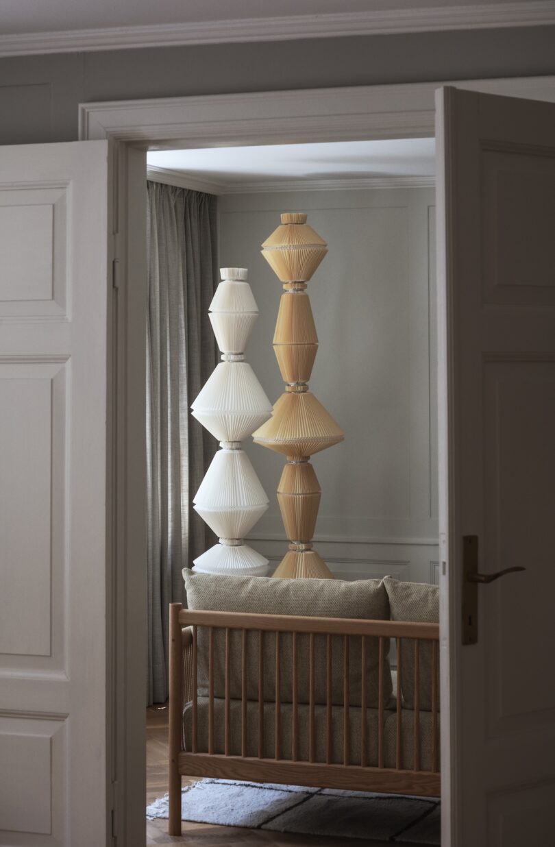 Two tall, geometric lamps with pleated shades, one white and one beige, stand behind a wooden sofa in a neutral-toned living room seen through an open door.