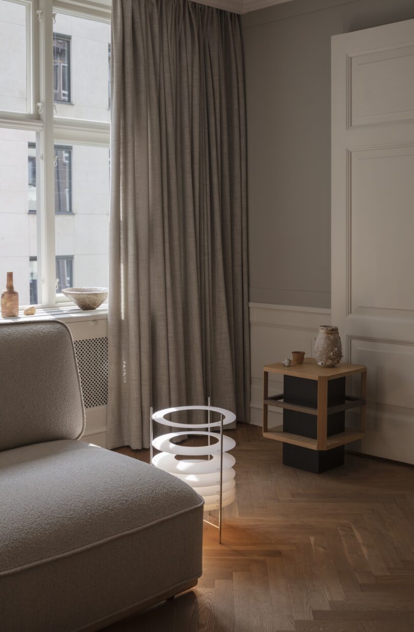 A modern living room with neutral tones, featuring a beige sofa, a round white floor lamp, a small side table with pottery, and floor-length curtains near a large window.
