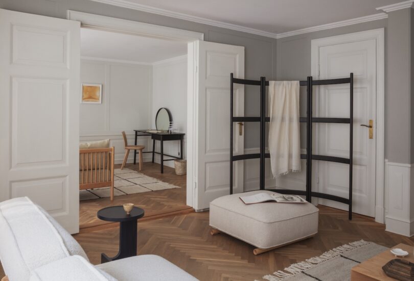 A modern living room with neutral tones features a beige ottoman, a black side table, a wooden room divider with a draped blanket, and an adjacent office area visible through open double doors.