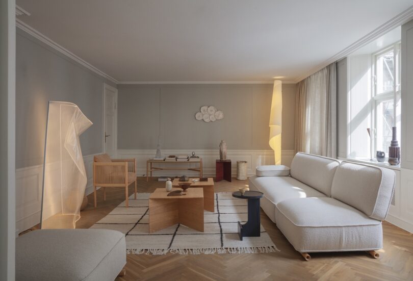 Minimalist living room with neutral-toned furniture, wooden accents, geometric coffee tables, a striped rug, and large windows letting in natural light.