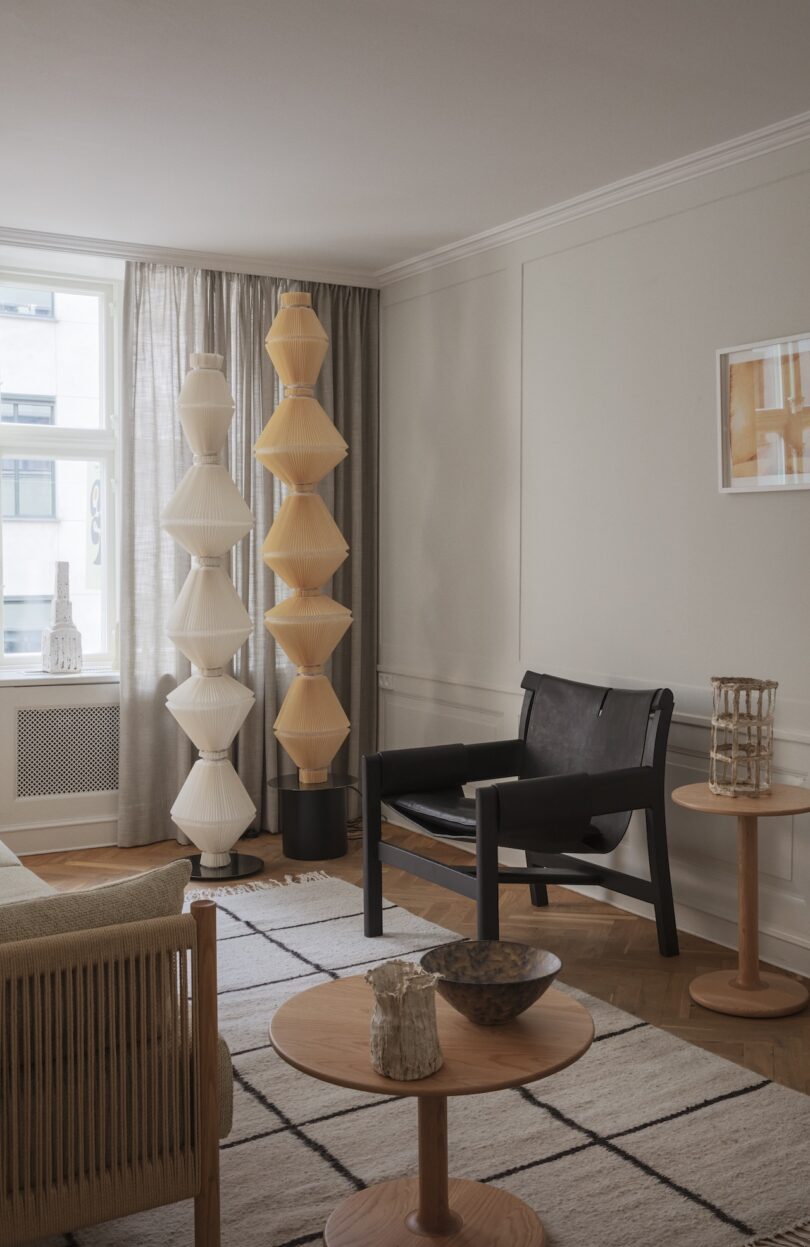 Minimalist living room with neutral tones, modern furniture, geometric rug, two sculptural floor lamps by the window, and decorative objects on wooden tables.