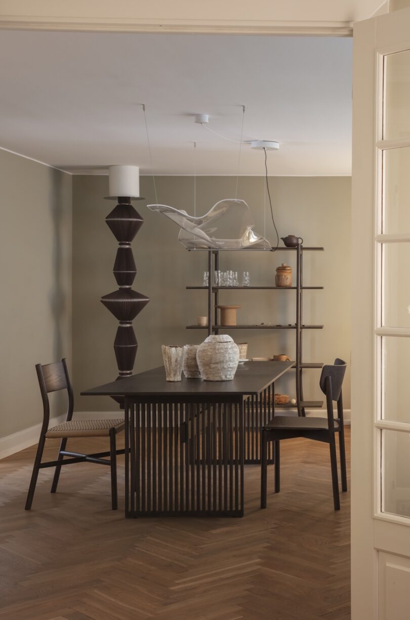Modern dining room with a dark wooden table, two chairs, decorative vases, a sculptural pendant light, and a shelving unit with pottery against beige walls and herringbone wood flooring.